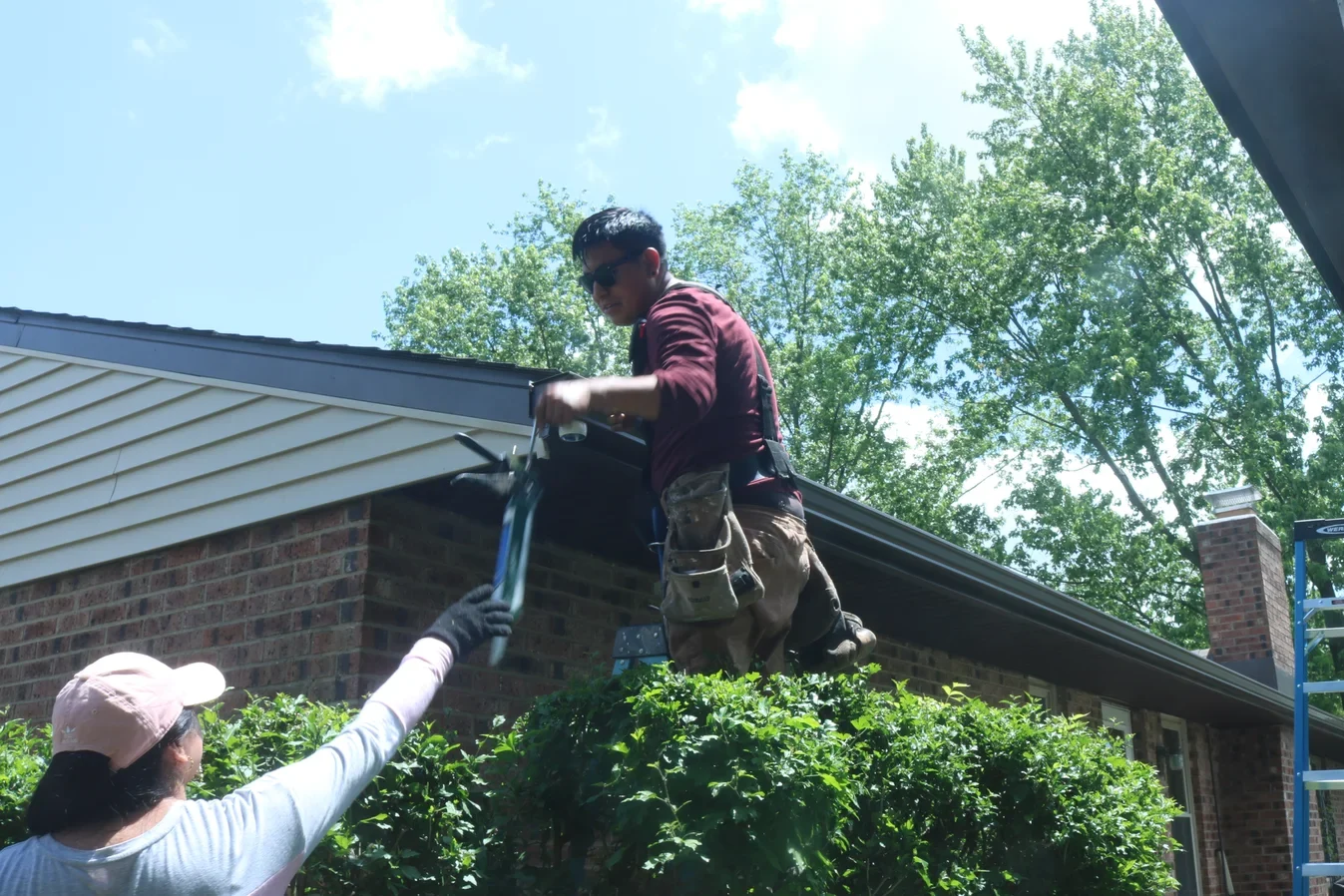 A worker standing on a ladder uses a saw on the roof of a house, possibly for gutter repair, while another person on the ground extends a hand for assistance.