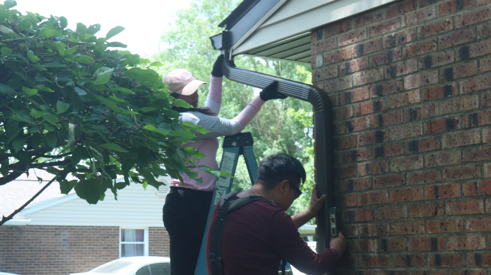 Two workers installing gutters on a brick house, one on a ladder and one handing tools
