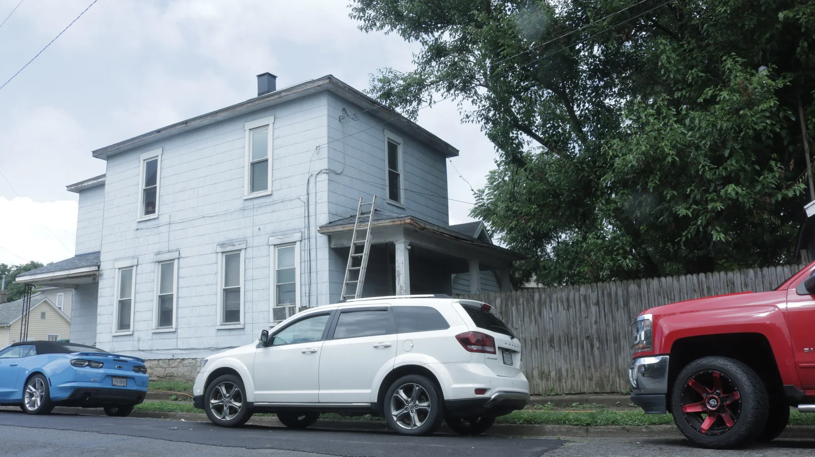 An old white house with a ladder leaning against it, a white SUV, and a red truck parked in front