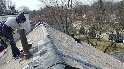 A worker in a hoodie and mask stands on a house roof, installing new shingles with tools for roof replacement, against the backdrop of a quiet residential area.