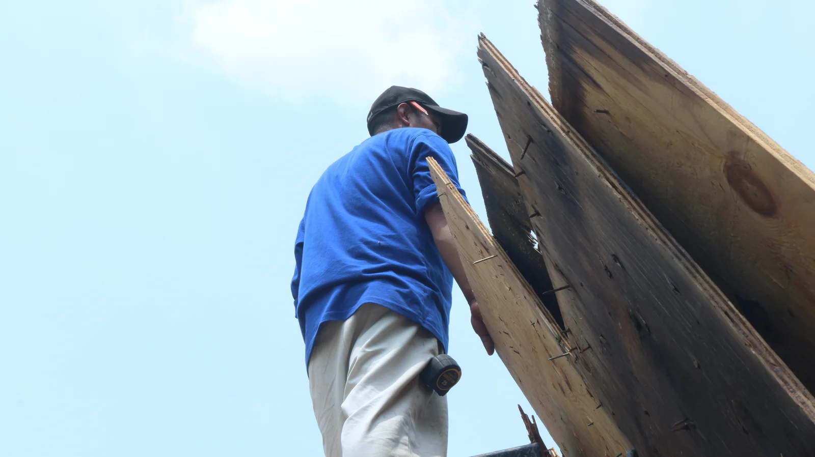 A worker in a blue shirt and white pants stands on a roof, viewed from below, holding a piece of plywood - roof inspection