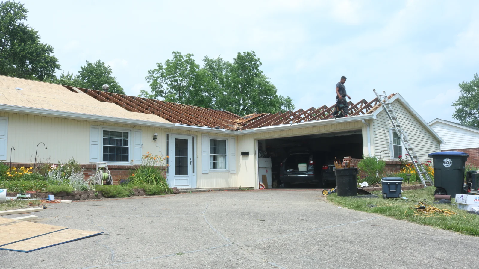Roof Repair : A house with its roof partially removed, showing exposed rafters, while a worker stands on the roof and another works on the ground
