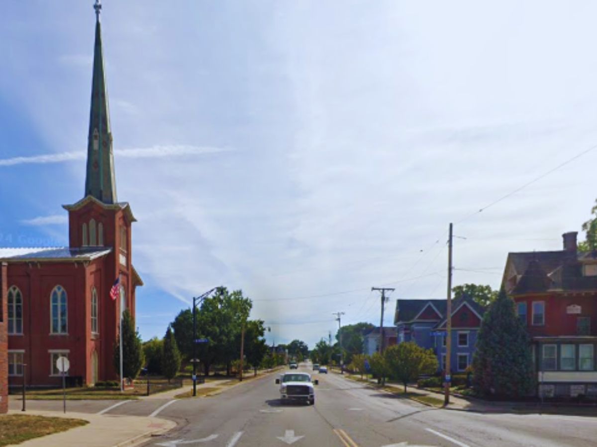 A vintage car cruises down 116 E Central Ave, Miamisburg, OH, with a steepled church on the left and quaint houses on the right.