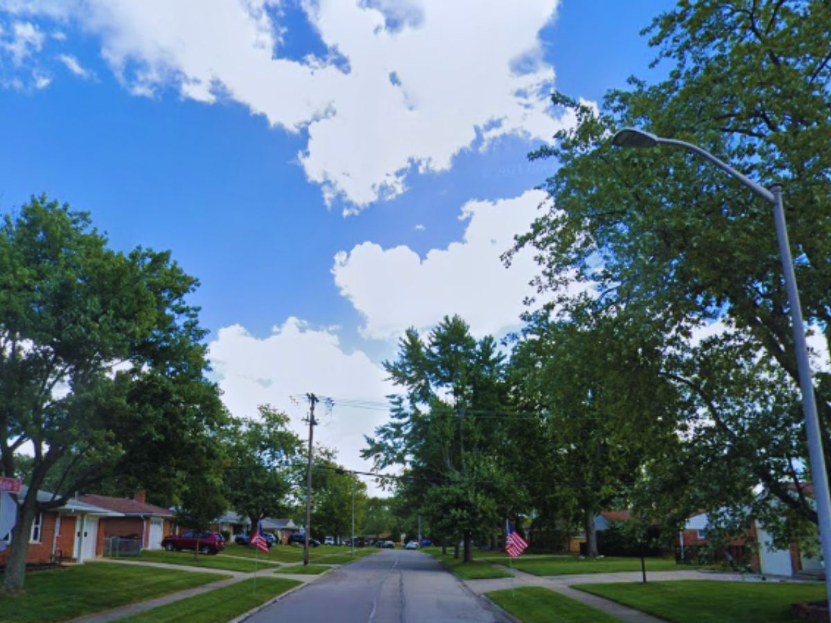 A sunny day on Kettering's 4200 Renwood Dr, with a tree-lined street, charming houses, blue skies, and scattered clouds.