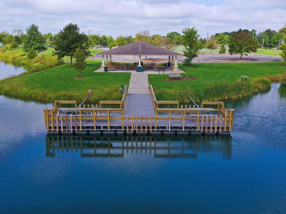 A wooden dock in Centerville, OH, leads to a pavilion with picnic tables, surrounded by lush grass and trees next to a pond under a cloudy sky.