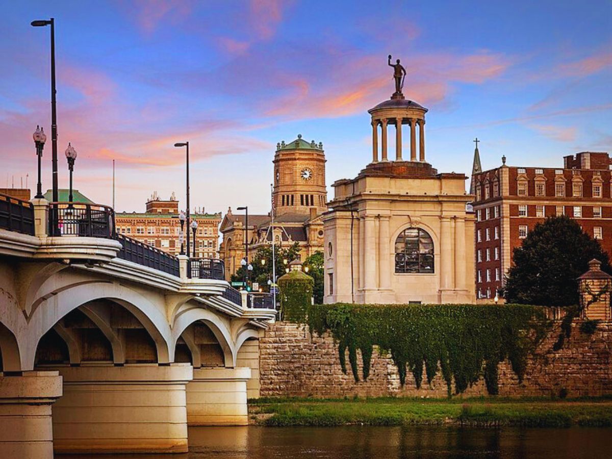 Bridge over river with a historic building featuring a statue and clock tower in the background during sunset, showcasing downtown Hamilton, OH.