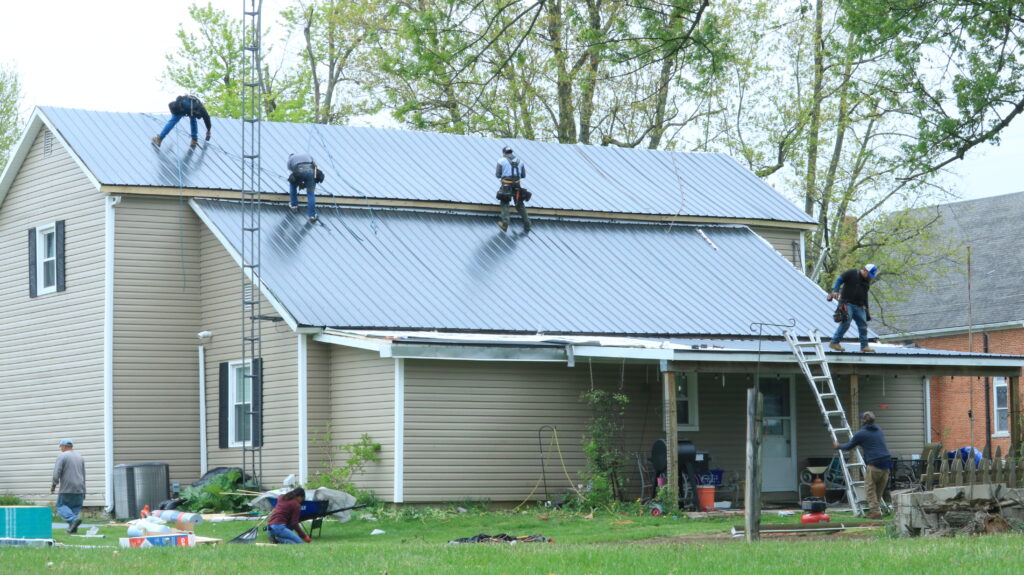 Workers from a top roofing service in Miami County repair a large metal roof on a two-story house, tools and materials scattered about.