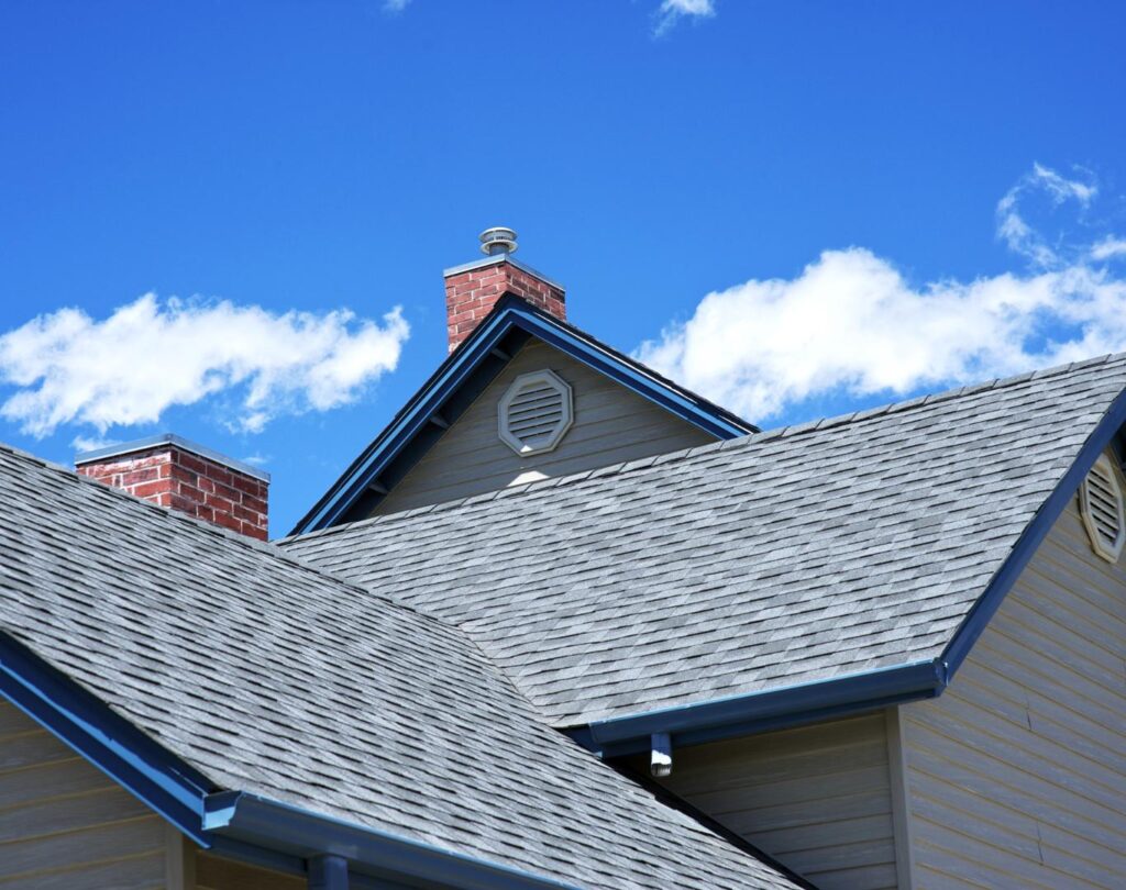 Close-up of house rooftops with gray shingles in Montgomery County, OH, set against a blue sky. Two red brick chimneys rise above the roofs.