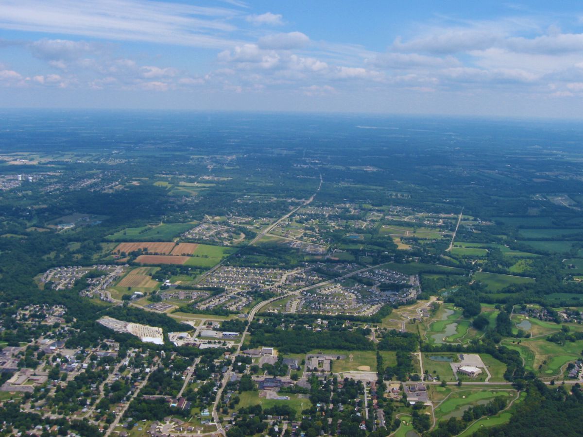 Aerial view of a sprawling suburban landscape in Warren County, OH, with roads, houses, and green fields under a blue sky with scattered clouds.