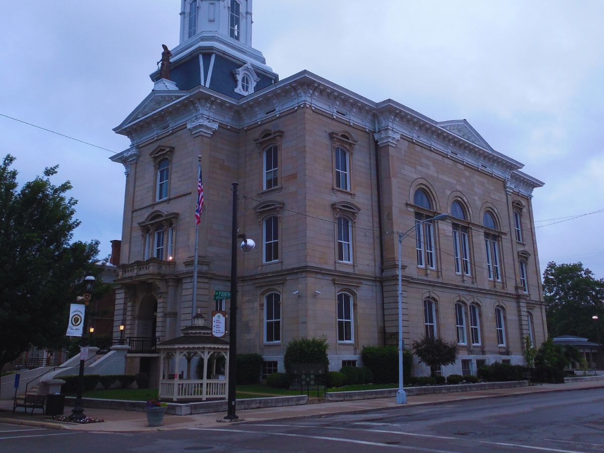 Historic Darke County brick courthouse with clock tower, nestled among trees on a quiet OH street corner. A U.S. flag flutters near the entrance.