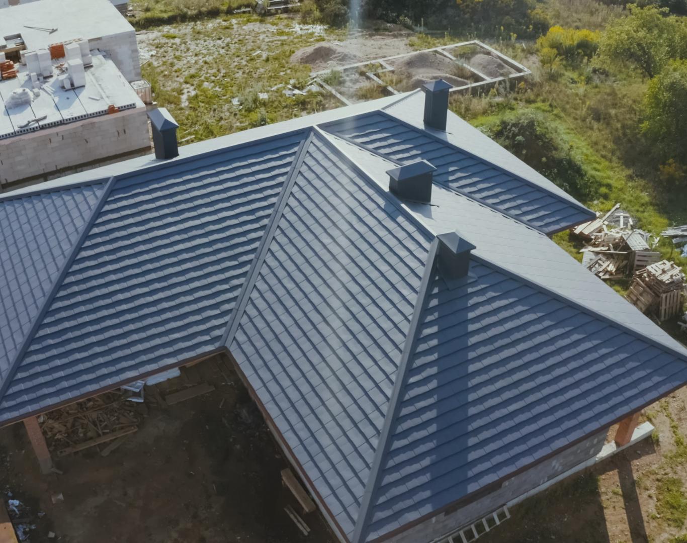 Aerial view of a gray multi-hipped roof under construction with chimneys, showcasing roofing services amid greenery and building materials.