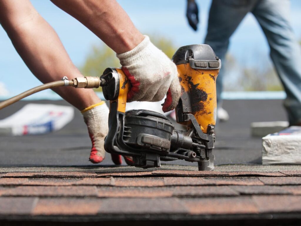 Person using a nail gun to install shingles on a roof, wearing gloves. A second person is partially visible in the background.