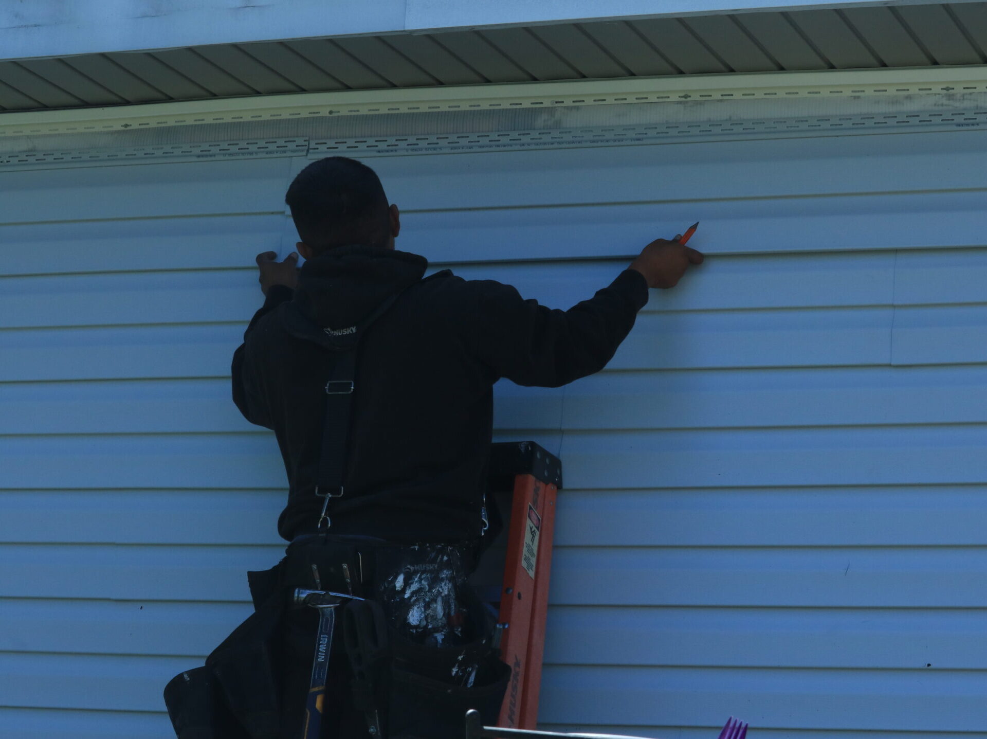 Person standing on a ladder, measuring and marking a blue house exterior for All-Side Roofing with a red tool.