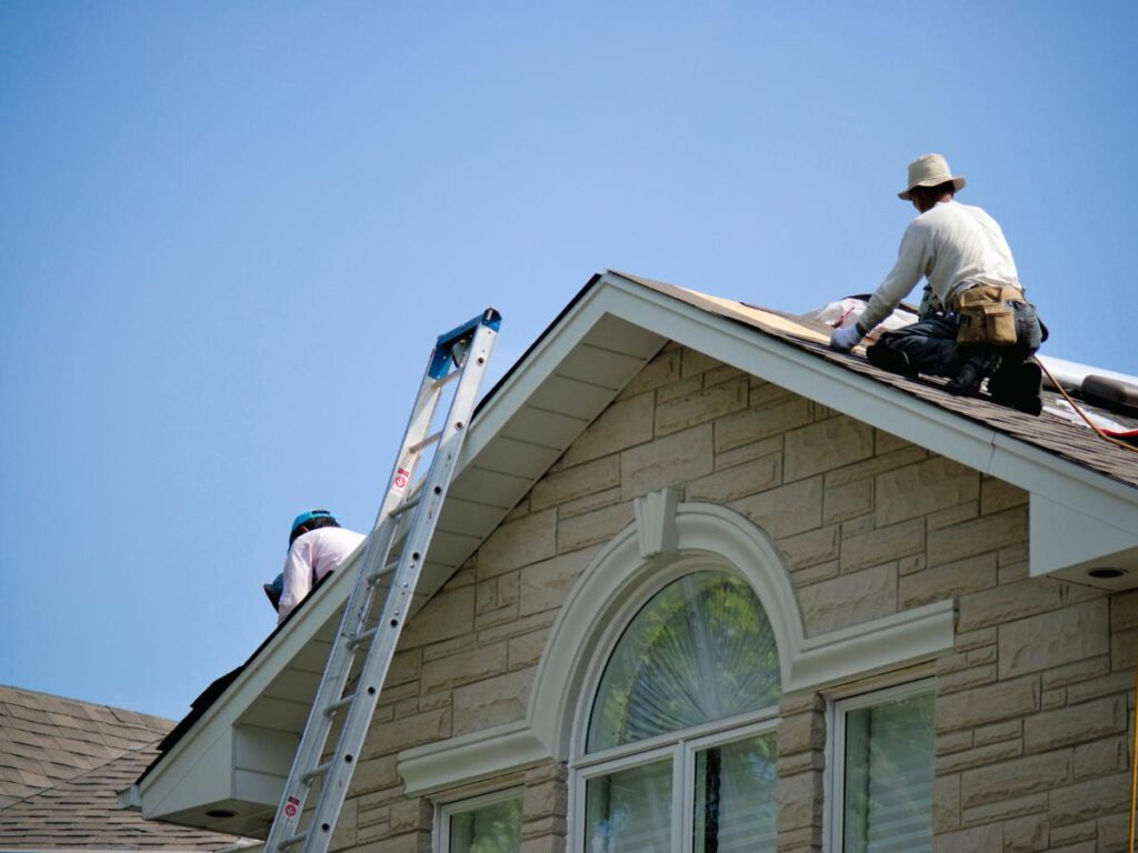 In OH, our roofing company in Darke County repairs a roof under a clear blue sky, with two workers in protective gear and a ladder against the building.