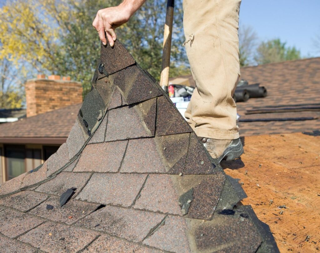 In Centerville, OH, a person removes old roof shingles with one hand on the shingle sheet and legs visible, set against a clear sky backdrop.