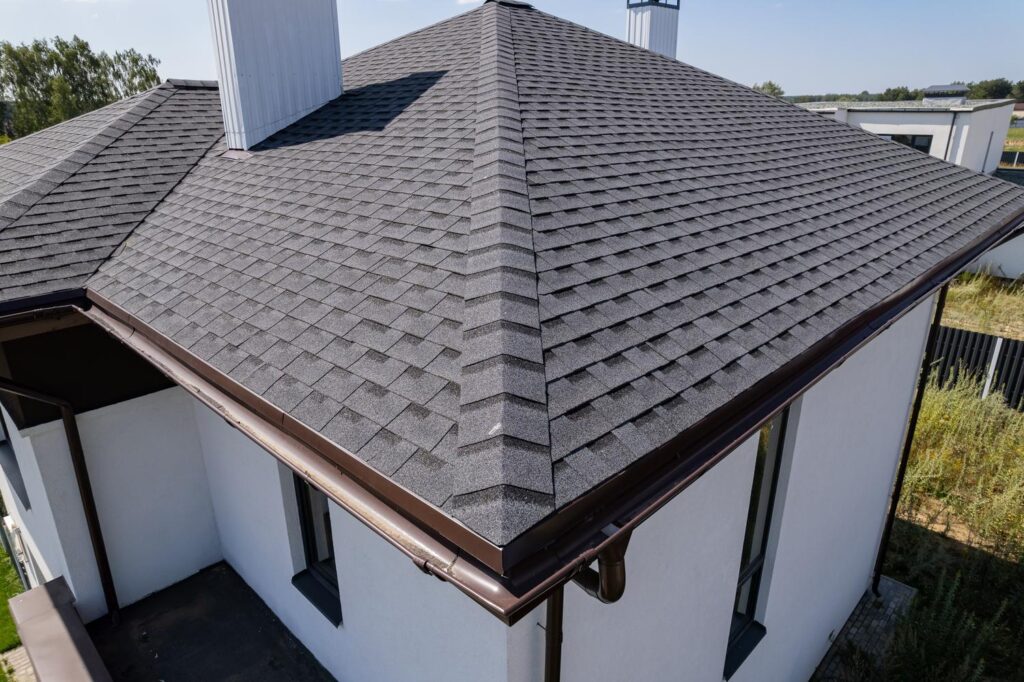A house with a dark gray shingle roof by a roofing company in Huber Heights, OH; white walls and brown gutters, surrounded by lush vegetation.