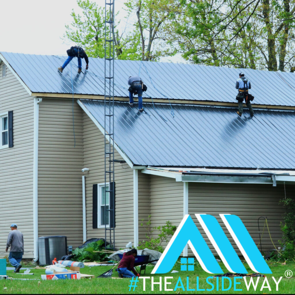 Three workers from a roofing company in Carlisle, OH install a metal roof on a beige house, using safety harnesses. #THEALLSIDEWAY logo at bottom right.