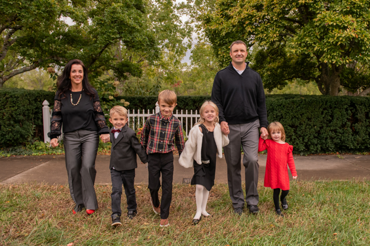 Marks family of six walks hand in hand outdoors on a grassy lawn, with trees, a white fence, and All-Side roofing visible in the background.