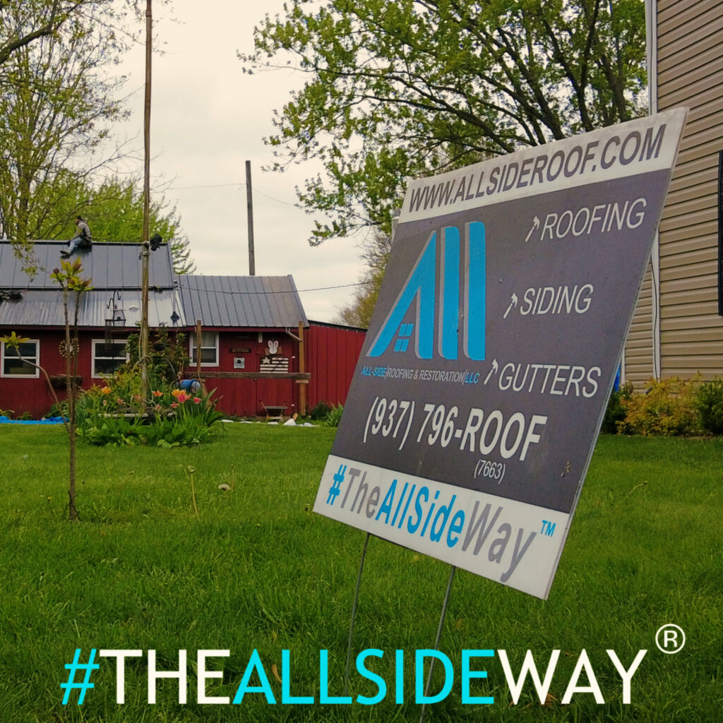 A yard sign in Kettering, OH advertises roofing, siding, and gutters while a crew works on roof repair in the background.