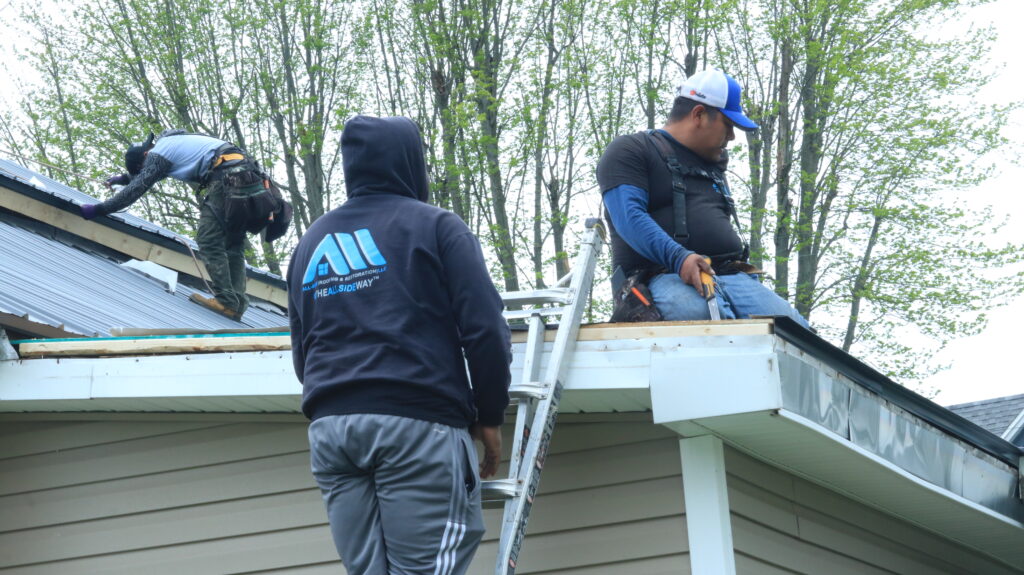 Three workers in safety gear perform roof repair on a house in Vandalia, OH, using ladders and tools amid trees on a cloudy day.