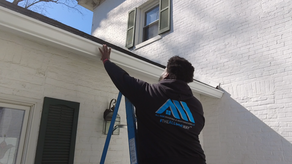 Person on a ladder inspects the roof gutter of a white brick house with green shutters in St Paris, OH for roof repair.