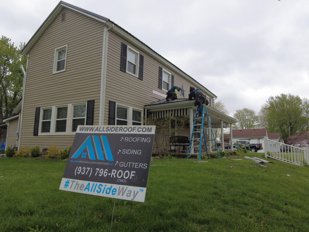 Two workers on ladders repair the front porch of a beige house in Springfield, OH. A yard sign displays Roof Repair company details and contact info.