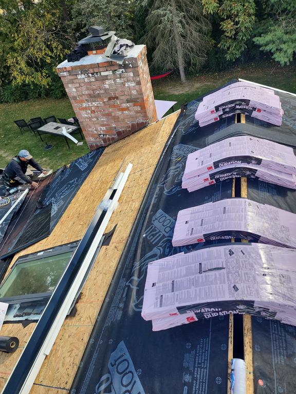 A person in Springboro, OH, works on a roof near a chimney during a roof replacement; trees and the yard are visible below.