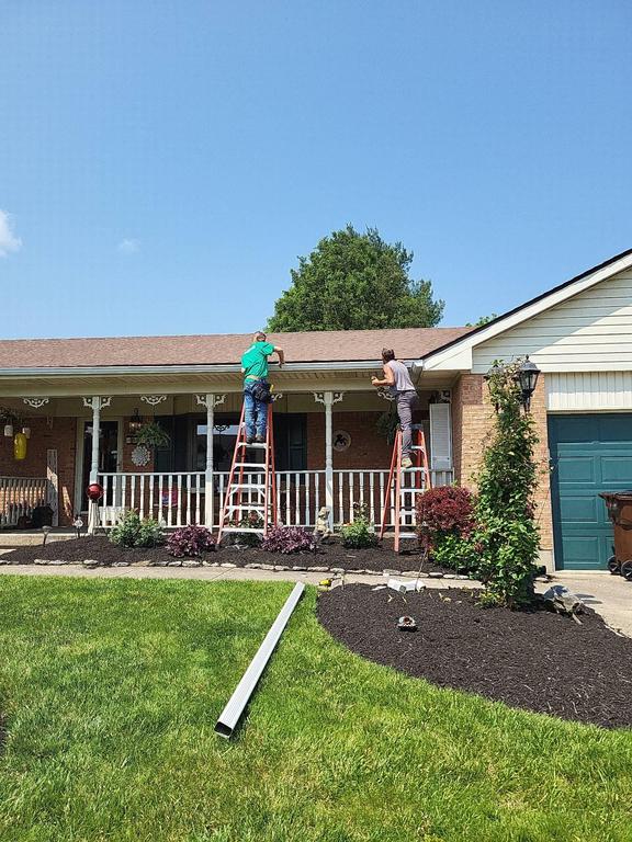Two people in Carlisle, OH work on a single-story brick house’s roof replacement, tools and materials scattered below on a sunny day.