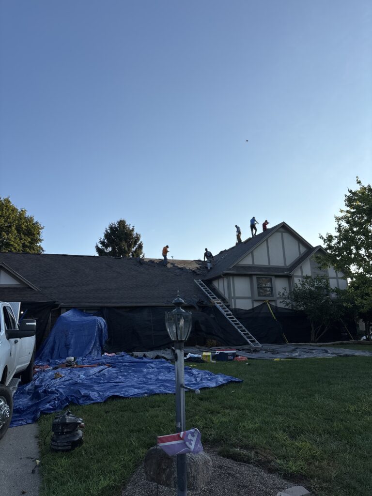 Roofing contractors perform a roof replacement on a house with protective tarps laid out on the ground below.