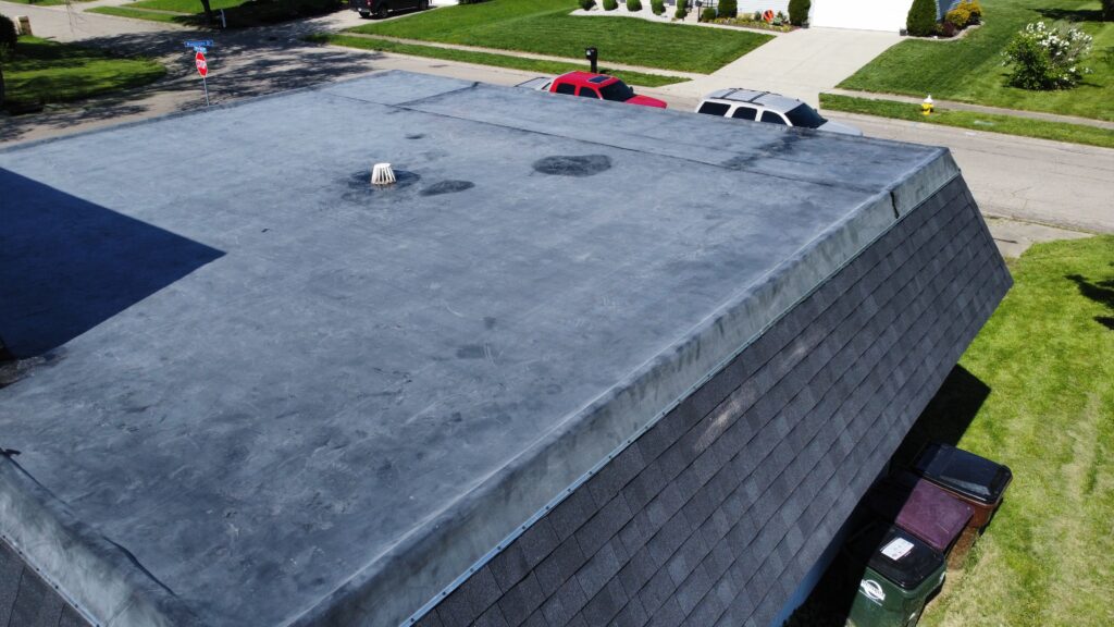 Aerial view of a flat rubber roof needing roof replacement, featuring a single vent pipe next to a sloped shingled roof and a residential street.